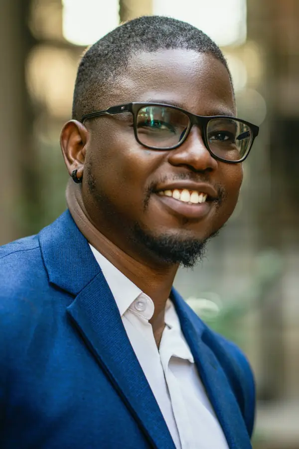 A smiling man with glasses in a blue blazer and white shirt, outdoors with soft green behind him.