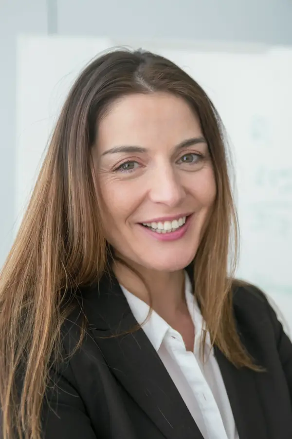 A smiling woman with long brown hair in a black blazer and white shirt, standing in an office.