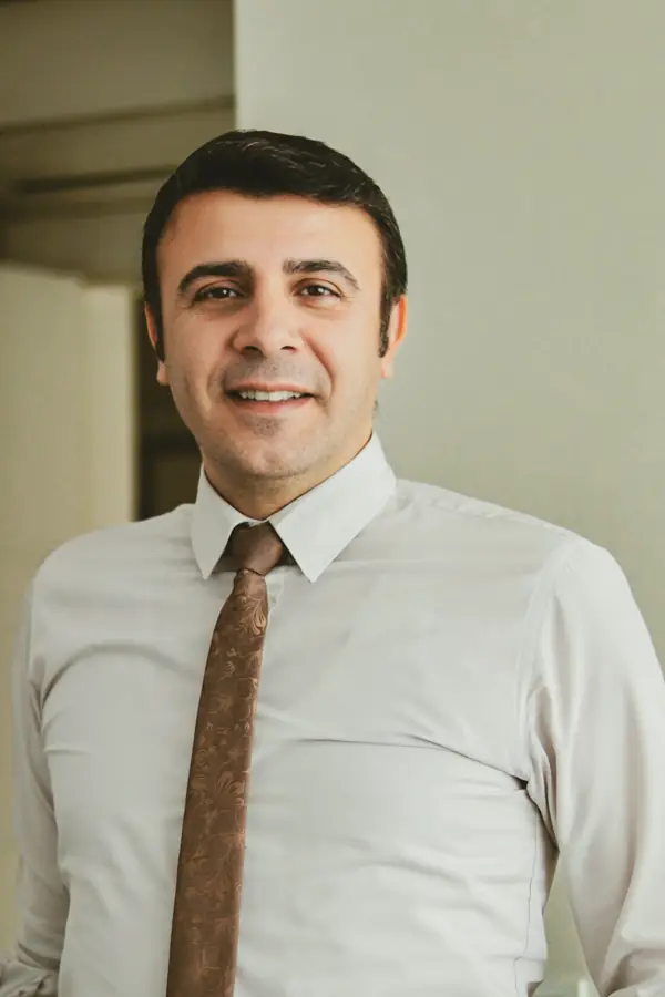 A smiling man in a white shirt and brown tie standing in a warm, sunlit hallway.