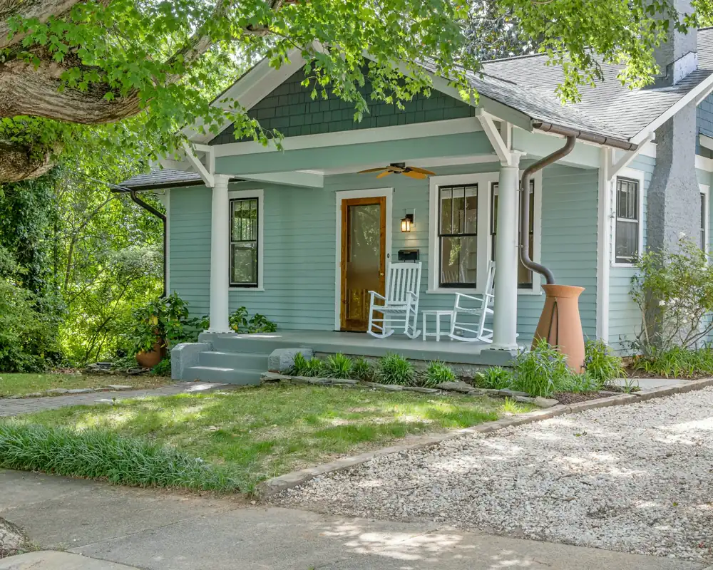 A pale blue craftsman bungalow with a deep front porch, square white columns, a wood door and two white rocking chairs.