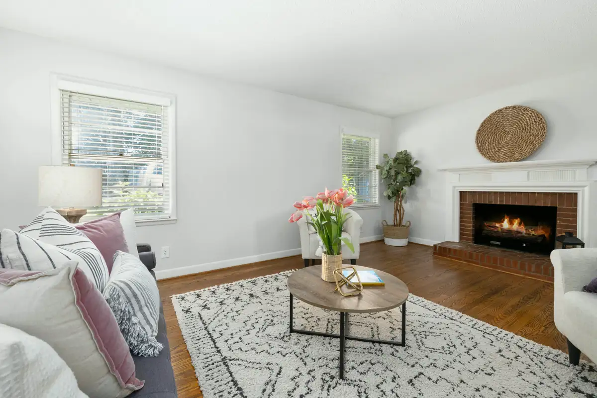 A staged family room with a corner fireplace, a white sofa with soft pink pillows, a round coffee table and a patterned rug on wood floors.