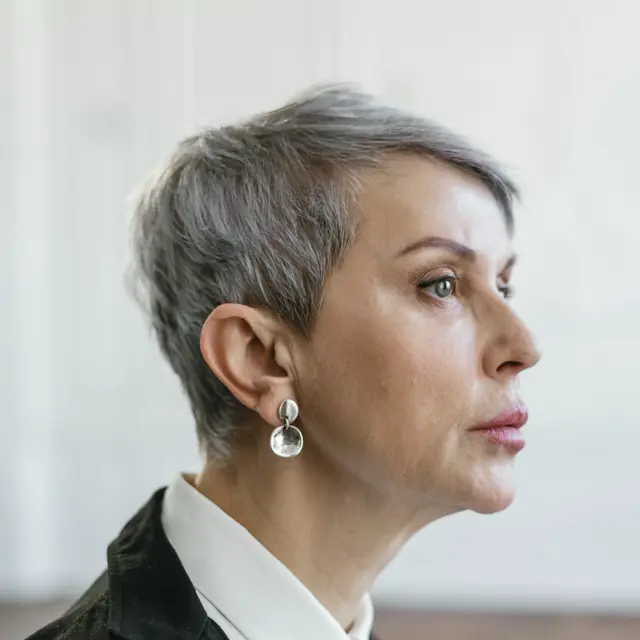 A woman with short silver hair and pearl earrings in a black blazer, seen in profile against a white paneled wall.
