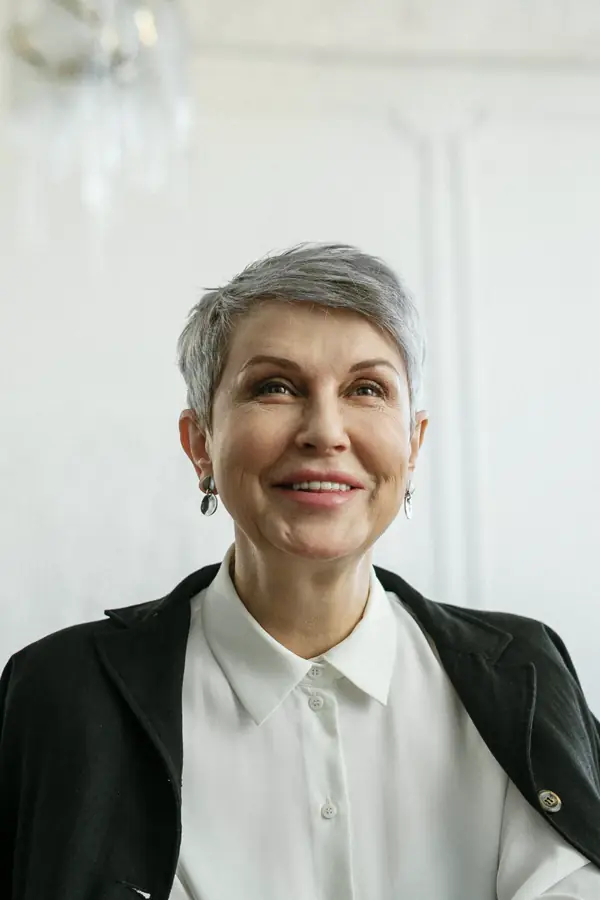 A smiling woman with short silver hair in a black blazer and white shirt, in a bright room with white paneled walls.