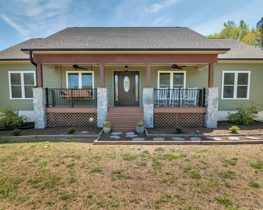 A green single-story house with a wide covered porch on stone pillars, a porch swing and rocking chairs, set back on a large lot.