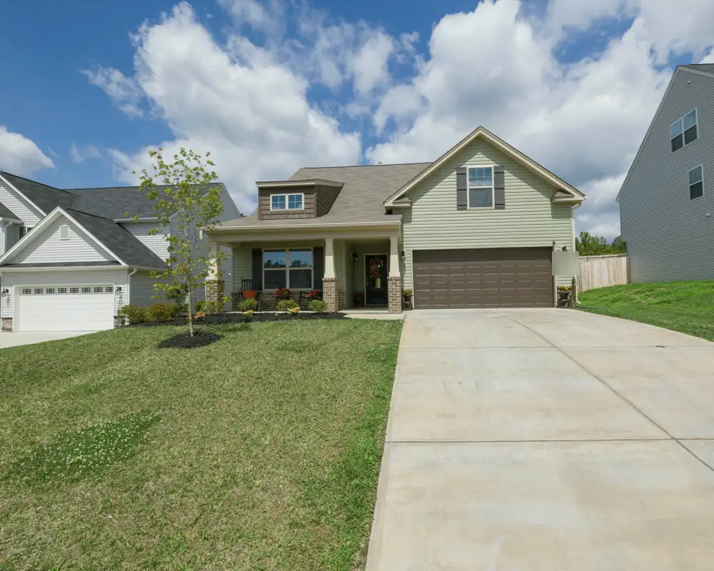 A sage green two-story house with a covered front porch, stone column bases and a brown two-car garage, behind a new lawn and a wide driveway.