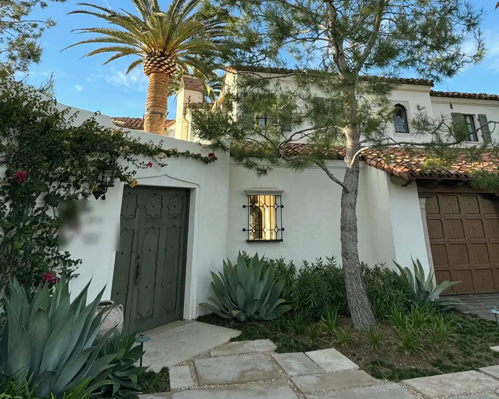 A white Spanish revival house with a tile roof, a green wooden door in the courtyard wall and a tall pine and palm in front.