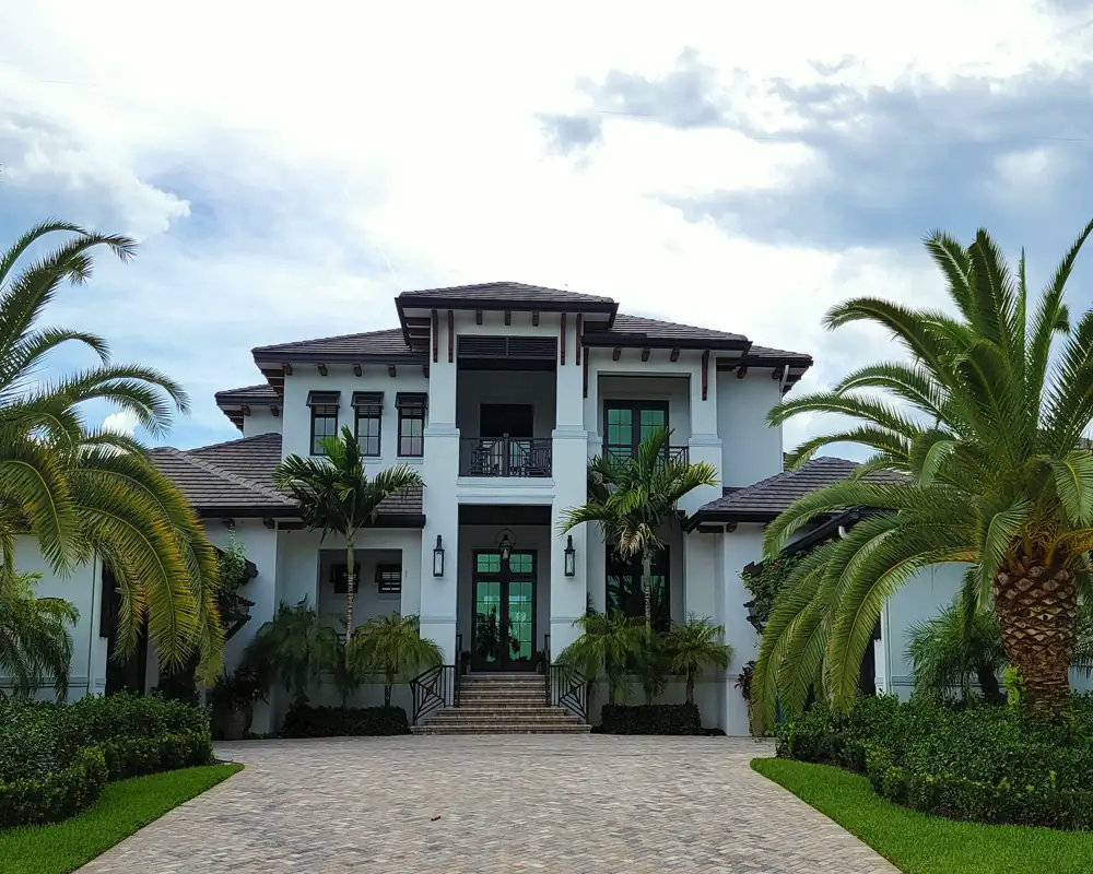 A white two-story house with dark trim, a second-floor balcony and a paver drive framed by two large palms.